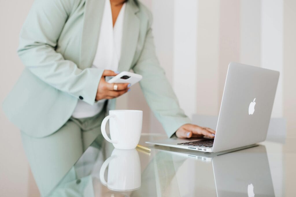 Professional woman in a mint green suit using a smartphone and Apple laptop at a modern office desk