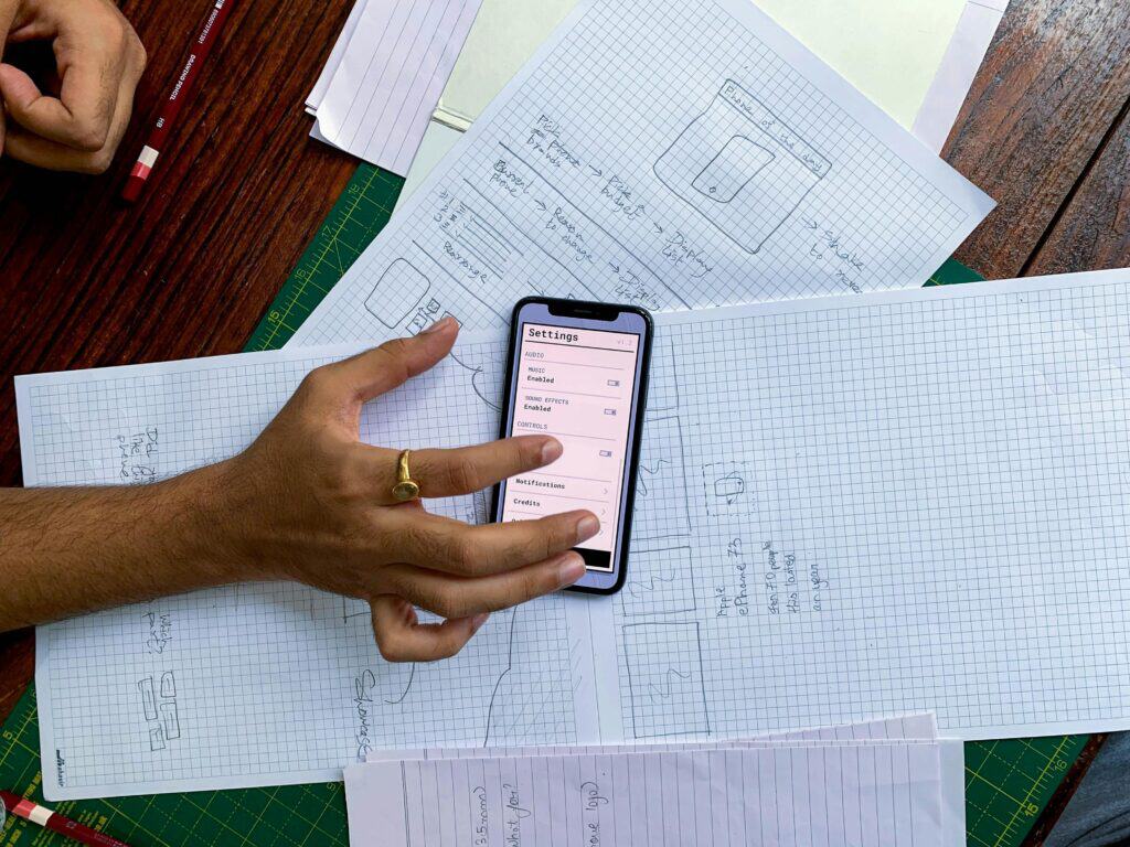 Developer reviewing hand-drawn mobile app wireframes and sketches alongside a prototype settings screen during the app planning stage