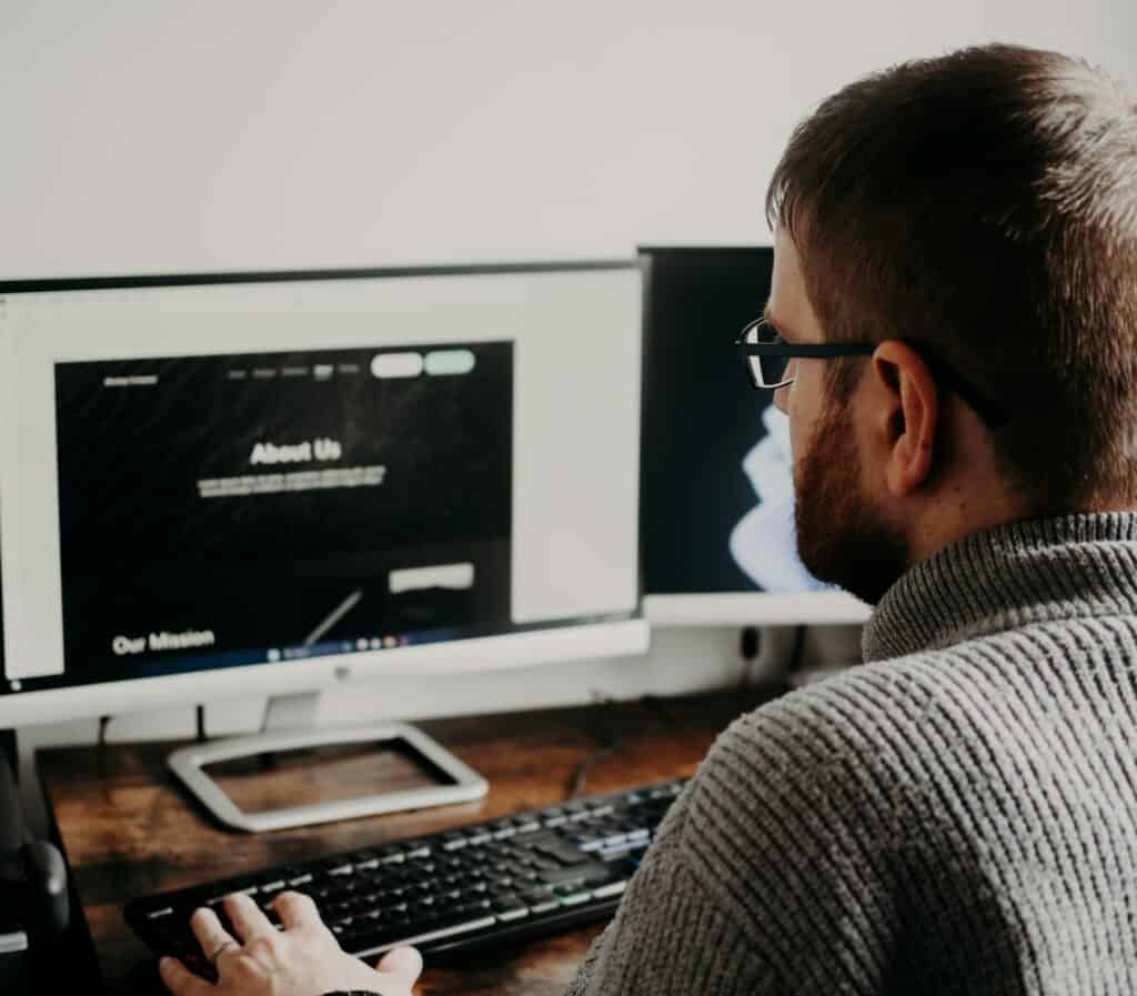 Web developer reviewing a website design on dual monitors at a desk, evaluating digital agency work in a home office setting